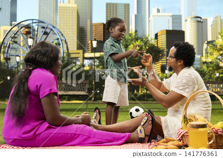 Young child smiling while playing rock paper scissors with father as mother watches during relaxed family picnic in sunny city park. Fun family time with playful kid on picnic blanket. 141776361