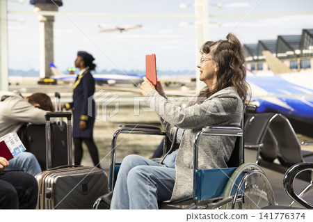 Joyful woman with impairment in wheelchair takes picture of flight details on screen, ready for flight departure. Elderly couple representing disability-friendly travel, mobility aid and freedom. Joyful woman with impairment in wheelchair takes picture of flight details on screen, ready for flight departure. Elderly couple representing disability-friendly travel, mobility aid and freedom. 141776374