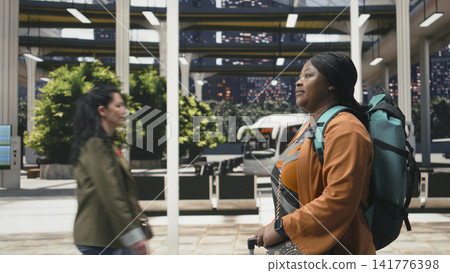 Traveler carries backpack and trolley through departure hall while heading toward next bus connection. Woman keeps personal bags close during long distance coach travel Traveler carries backpack and trolley through departure hall while heading toward next bus connection. Woman keeps personal bags close during long distance coach travel 141776398