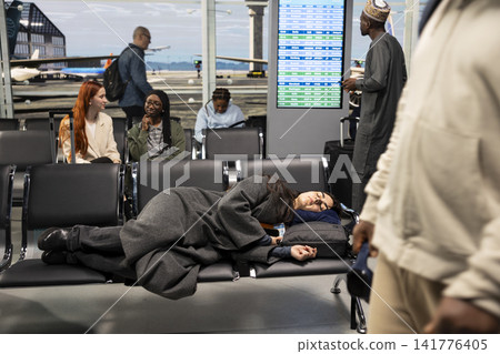 Jet lagged woman sleeping on airport bench while waiting for flight, passing time. Bored girl taking nap on departure hub chairs while waiting for plane taking her to travel destination 141776405