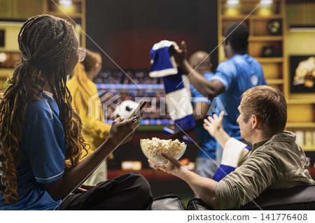 Woman watching live soccer match in TV with phone in hand while seated on couch. Football fans enjoying game broadcast on screen, enjoying snacks snack on sofa Woman watching live soccer match in TV with phone in hand while seated on couch. Football fans enjoying game broadcast on screen, enjoying snacks snack on sofa 141776408