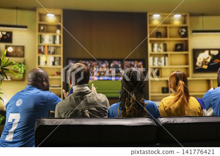 Football fans in team jerseys watch match on TV from living room sofa. Rear view of soccer supporters group wearing kits in team colors following television screen 141776421