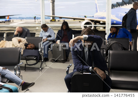 Tired CEO man in airport terminal lounge trying to rest during delayed flight business trip. Exhausted businessman sleeping on trolley handle, waiting for canceled flights to resume Tired CEO man in airport terminal lounge trying to rest during delayed flight business trip. Exhausted businessman sleeping on trolley handle, waiting for canceled flights to resume 141776455