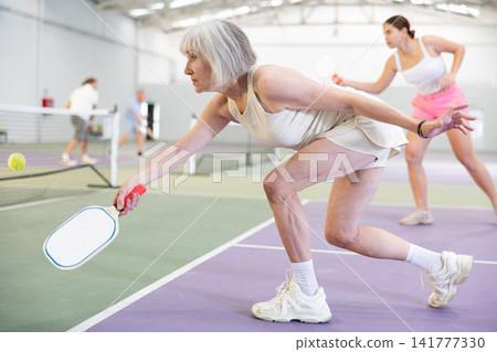 Elderly woman bouncing ball in pickleball game on court with racket Elderly woman bouncing ball in pickleball game on court with racket 141777330