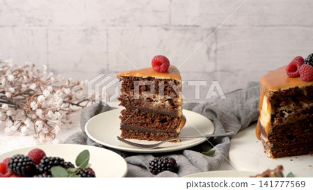 Chocolate cake slice with caramel frosting and a raspberry on top, surrounded by fresh berries and a floral arrangement, creating a cozy dessert scene Chocolate cake slice with caramel frosting and a raspberry on top, surrounded by fresh berries and a floral arrangement, creating a cozy dessert scene 141777569