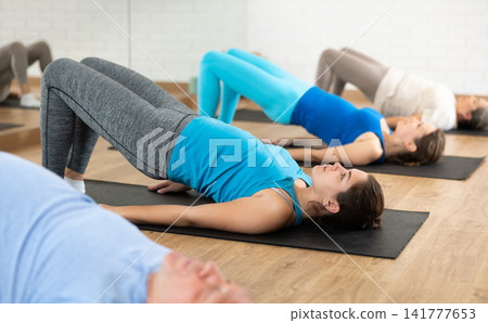 Class of male and women of different ages in active wear doing buttock bridge exercise during Pilates training together in gym Class of male and women of different ages in active wear doing buttock bridge exercise during Pilates training together in gym 141777653