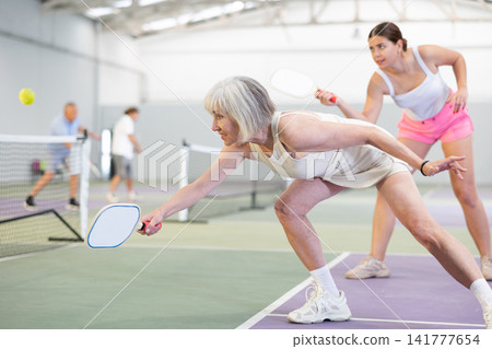 Elderly woman bouncing ball in pickleball game on court with racket Elderly woman bouncing ball in pickleball game on court with racket 141777654