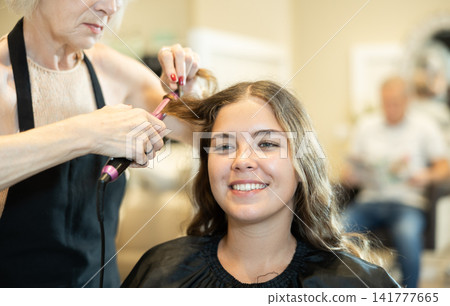 In beauty salon, cropped elderly female hairdresser makes styling curls for young girl client In beauty salon, cropped elderly female hairdresser makes styling curls for young girl client 141777665