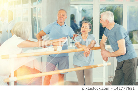 Warm-up sessions, mature women and men doing ballet exercise and talking, standing beside ballet barre in hall Warm-up sessions, mature women and men doing ballet exercise and talking, standing beside ballet barre in hall 141777681