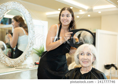 Young woman hairdresser holds mirror behind senior female client and shows result of work. Young woman hairdresser holds mirror behind senior female client and shows result of work. 141777746