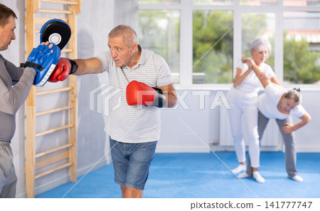 Old man training boxing kicks on punch mitts held by instructor Old man training boxing kicks on punch mitts held by instructor 141777747