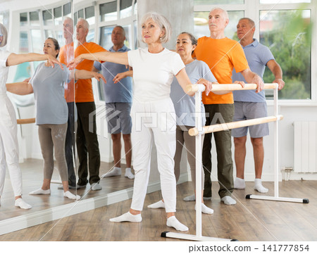 Pensioners of different ages exercise near ballet barre at group training session in two position Pensioners of different ages exercise near ballet barre at group training session in two position 141777854