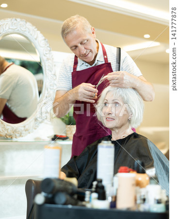 Experienced senior man master makes performs haircut for mature woman with scissors Experienced senior man master makes performs haircut for mature woman with scissors 141777878