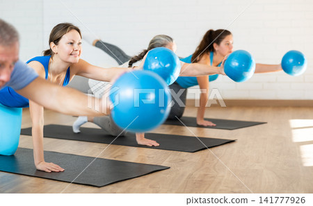 Class of male and women of different ages in active wear working out with mini balls during Pilates training together in rehabilitation center Class of male and women of different ages in active wear working out with mini balls during Pilates training together in rehabilitation center 141777926
