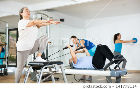 Aged woman practicing pilates on pedal fitness chair in sports hall Aged woman practicing pilates on pedal fitness chair in sports hall 141778114