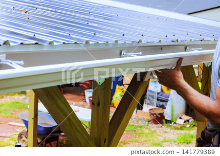 Worker is attaching metal roofing gutter panels on wooden frame in an outdoor setting tools equipment Worker is attaching metal roofing gutter panels on wooden frame in an outdoor setting tools equipment 141779389