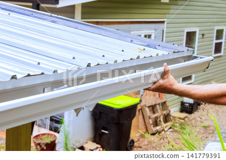 Worker installs metal roof gutter on house at bright light, residential area in background. Worker installs metal roof gutter on house at bright light, residential area in background. 141779391