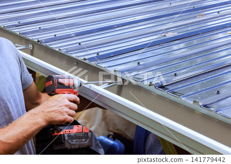 Builder uses drill to attach metal roof gutter on structure in residential location Builder uses drill to attach metal roof gutter on structure in residential location 141779442
