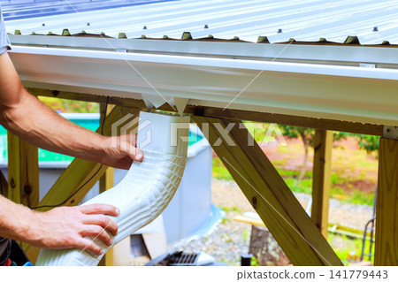 Handyman works on attaching rain gutter to roof of house it is setting is backyard. Handyman works on attaching rain gutter to roof of house it is setting is backyard. 141779443