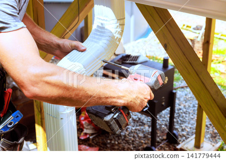 Worker holds piece of gutter while using drill to secure it work takes place at residential area. Worker holds piece of gutter while using drill to secure it work takes place at residential area. 141779444