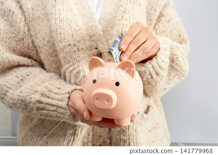 Heating season. Person inserts cash into piggy bank wearing cozy sweater Heating season. Person inserts cash into piggy bank wearing cozy sweater 141779968