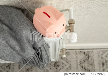 Heating season. Piggy bank rests on top of radiator while cozy blanket hangs nearby Heating season. Piggy bank rests on top of radiator while cozy blanket hangs nearby 141779972