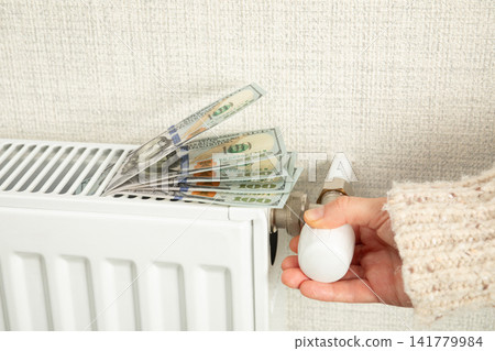 Heating season. Homeowner turns thermostat while cash lies on radiator Heating season. Homeowner turns thermostat while cash lies on radiator 141779984