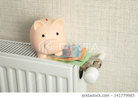 Heating season. Piggy bank sits atop radiator next to bills related to heating season Heating season. Piggy bank sits atop radiator next to bills related to heating season 141779985