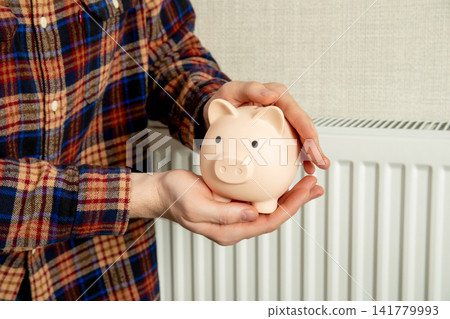 Heating season. Person holds piggy bank in front of radiator to save on heating season expenses Heating season. Person holds piggy bank in front of radiator to save on heating season expenses 141779993