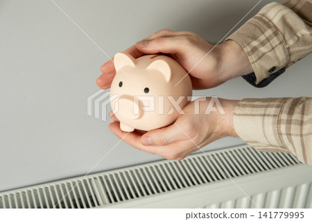 Heating season. Individual holds piggy bank near radiator while considering heating costs Heating season. Individual holds piggy bank near radiator while considering heating costs 141779995
