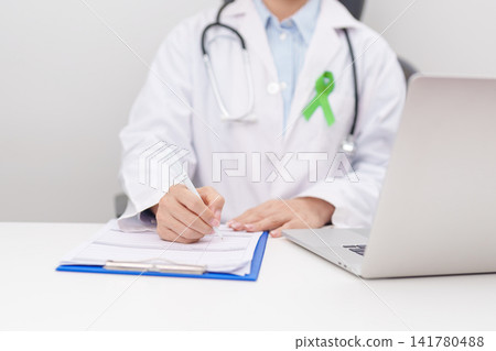 doctor using laptop and report notes on notebook. lime Green Ribbon for concept of May Celiac Awareness month, Lyme disease, September Lymphoma, Mental Health Awareness month and October Liver cancer doctor using laptop and report notes on notebook. lime Green Ribbon for concept of May Celiac Awareness month, Lyme disease, September Lymphoma, Mental Health Awareness month and October Liver cancer 141780488