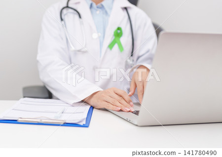 doctor using laptop and report notes on notebook. lime Green Ribbon for concept of May Celiac Awareness month, Lyme disease, September Lymphoma, Mental Health Awareness month and October Liver cancer doctor using laptop and report notes on notebook. lime Green Ribbon for concept of May Celiac Awareness month, Lyme disease, September Lymphoma, Mental Health Awareness month and October Liver cancer 141780490