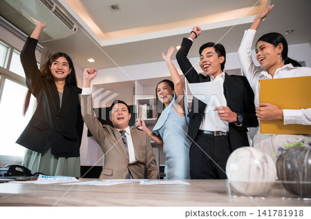 Excited business team celebrating success in modern office and raising hands with joyful. Asian professionals in formal wears expressing triumph after achieving goals or closing deal. Excited business team celebrating success in modern office and raising hands with joyful. Asian professionals in formal wears expressing triumph after achieving goals or closing deal. 141781918