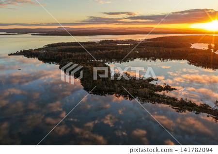 Aerial view of a sunset sky reflected across the calm waters of Pipers Bay, creating a peaceful coastal scene with soft colours, pastel tones, glassy water and a tranquil natural atmosphere. Aerial view of a sunset sky reflected across the calm waters of Pipers Bay, creating a peaceful coastal scene with soft colours, pastel tones, glassy water and a tranquil natural atmosphere. 141782094