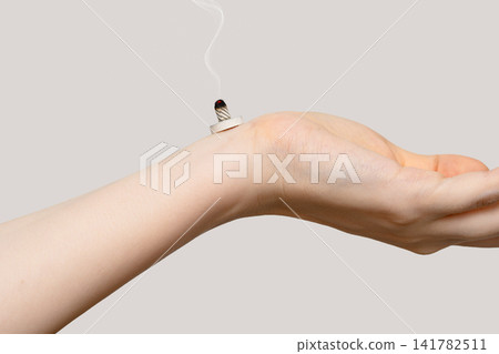 A burning moxa cone is emitting smoke near a woman's wrist. A burning moxa cone is emitting smoke near a woman's wrist. 141782511