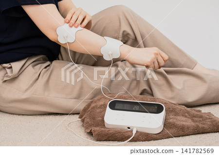 A woman uses a low-frequency therapy device with electrode pads attached to her arm. A woman uses a low-frequency therapy device with electrode pads attached to her arm. 141782569