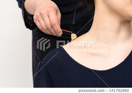 A hand applying incense to a moxa moxibustion treatment at the base of a woman's neck. A hand applying incense to a moxa moxibustion treatment at the base of a woman's neck. 141782660