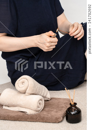 A woman lights incense in front of a towel and a small bottle of fragrance. A woman lights incense in front of a towel and a small bottle of fragrance. 141782692
