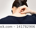 A woman's back view as she supports a moxa cone on the nape of her neck with her fingertips. 141782906