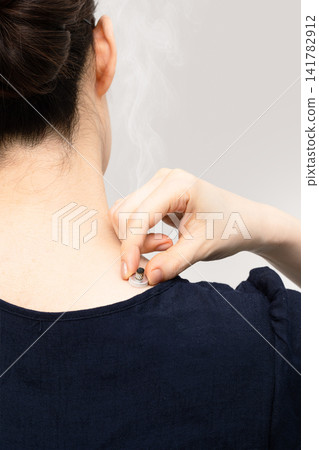 A woman with smoke rising from a moxa cone at the base of her neck and shoulder. A woman with smoke rising from a moxa cone at the base of her neck and shoulder. 141782912