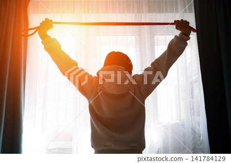 Person silhouette raising arms with a resistance band, stretching in front of a bright window, preparing for a morning fitness routine and physical exercise Person silhouette raising arms with a resistance band, stretching in front of a bright window, preparing for a morning fitness routine and physical exercise 141784129