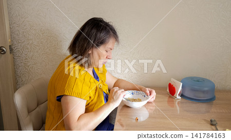 Woman in a yellow cardigan sitting at a wooden table, enjoying a bowl of cereal for a healthy breakfast in her home, focusing on nutrition and daily routine 141784163