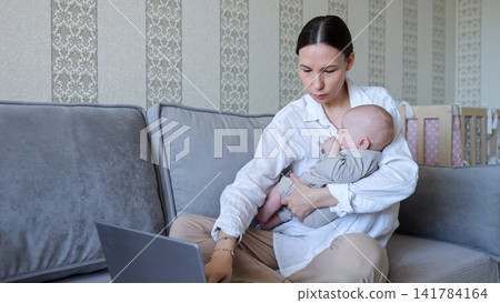 Young mother multitasking working on laptop holding baby Young mother multitasking working on laptop holding baby 141784164