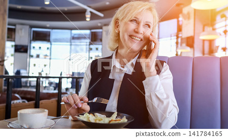 Caucasian Elderly Businesswoman Smiling On Phone While Eating Salad And Drinking Coffee At Sunlit Cafe Booth, Holding Pen And Jotting Notes, Relaxed Professional Energy, Casual Networking Caucasian Elderly Businesswoman Smiling On Phone While Eating Salad And Drinking Coffee At Sunlit Cafe Booth, Holding Pen And Jotting Notes, Relaxed Professional Energy, Casual Networking 141784165
