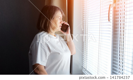 Senior woman in a white t shirt talking on a smartphone, looking out through horizontal blinds, sunlight streaming in, representing communication, solitude, and connection at home Senior woman in a white t shirt talking on a smartphone, looking out through horizontal blinds, sunlight streaming in, representing communication, solitude, and connection at home 141784237