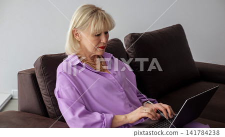 Senior woman with blonde hair wearing a purple shirt sitting comfortably on a brown sofa, actively typing on a laptop, engaging in online activity, remote work, or telecommuting at home 141784238