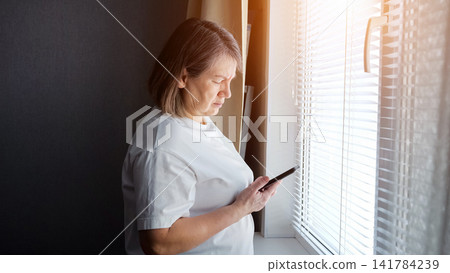 Senior woman standing near a sunlit window with blinds, looking at her smartphone with a serious expression, connecting with technology and the digital world from home Senior woman standing near a sunlit window with blinds, looking at her smartphone with a serious expression, connecting with technology and the digital world from home 141784239