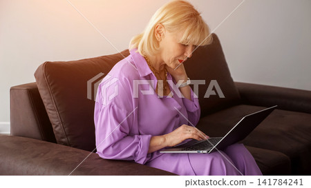 Senior woman relaxing on a couch, embracing modern technology by typing on a laptop, connecting to the internet, and engaging in digital activities at home Senior woman relaxing on a couch, embracing modern technology by typing on a laptop, connecting to the internet, and engaging in digital activities at home 141784241