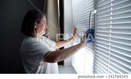 Woman carefully wiping dust off white horizontal window blinds using a blue microfiber cloth, performing a domestic chore to maintain a clean home environment Woman carefully wiping dust off white horizontal window blinds using a blue microfiber cloth, performing a domestic chore to maintain a clean home environment 141784243