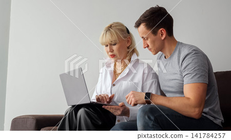 Mature woman and young man sit on a couch in a home office, collaborating over a laptop, mentoring and learning together, demonstrating intergenerational digital literacy and support 141784257